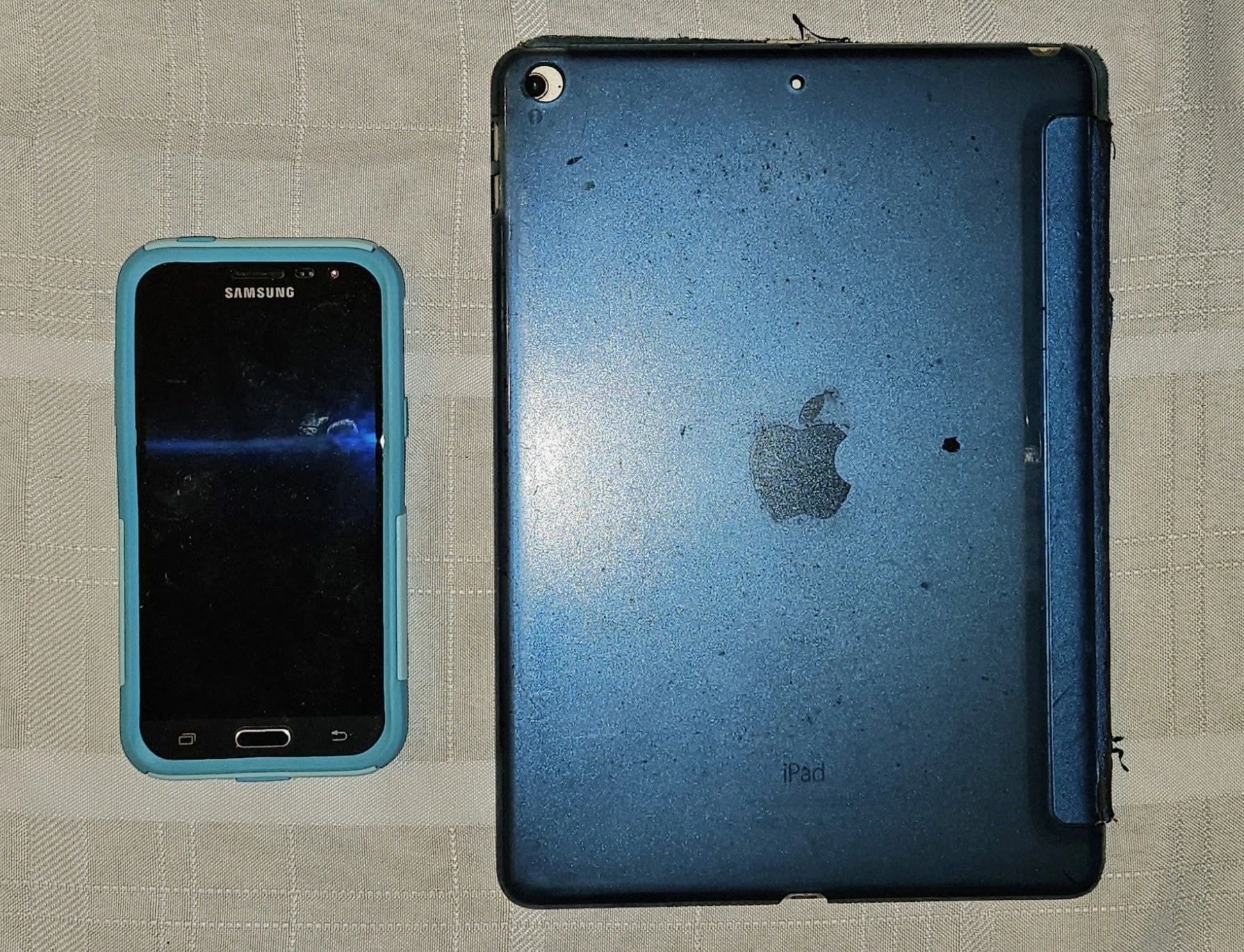 Samsung smartphone and blue Apple iPad placed side by side on a cloth surface.
